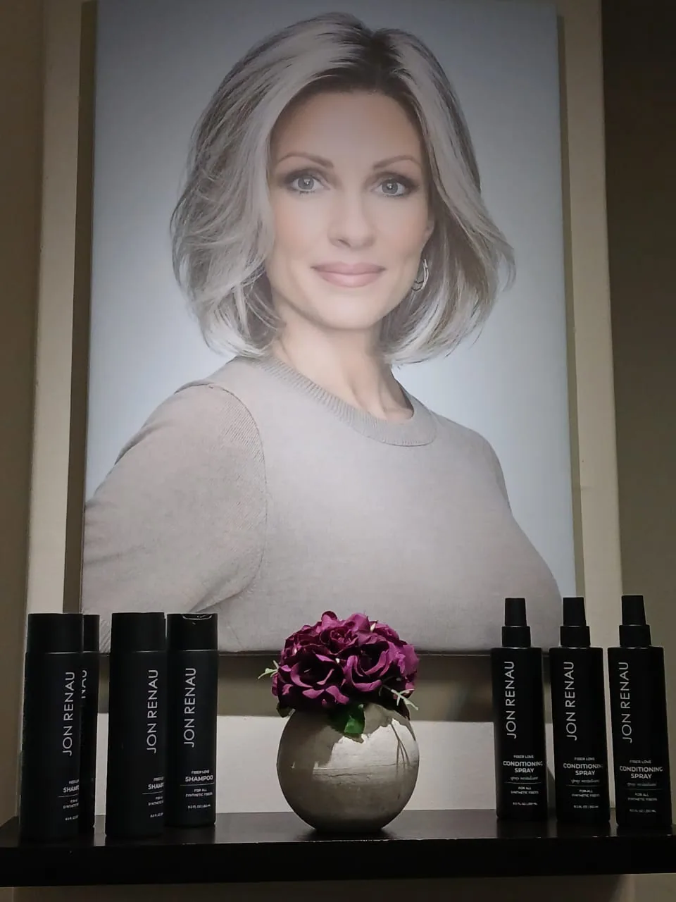 A silver-grey bob hairstyle poster above Jon Renau wig care products and purple flowers